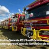 Linha de caminhões de bombeiros vermelhos com estruturas amarelas alinhados em um campo, sob céu azul; legenda informa investimento federal de R$24 milhões na Bahia para viaturas e equipamentos.